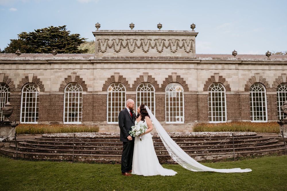 A couple in wedding attire poses outside a grand stone building with arched windows and steps.