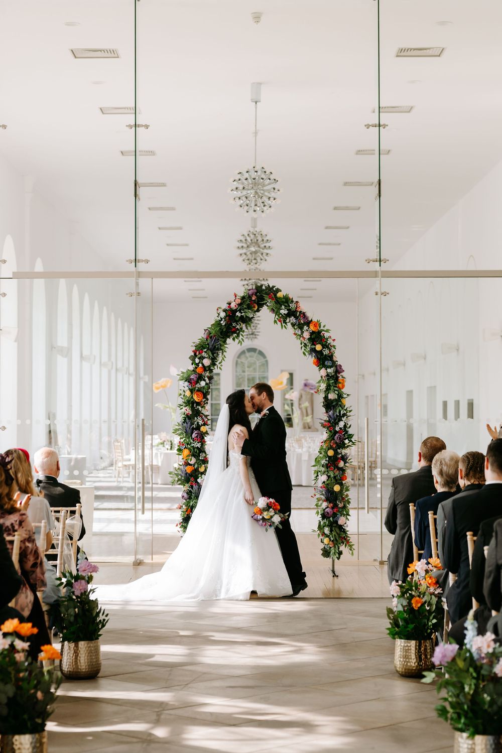 A wedding ceremony in a bright white venue with a floral arch, chandeliers, and guests seated along the aisle.