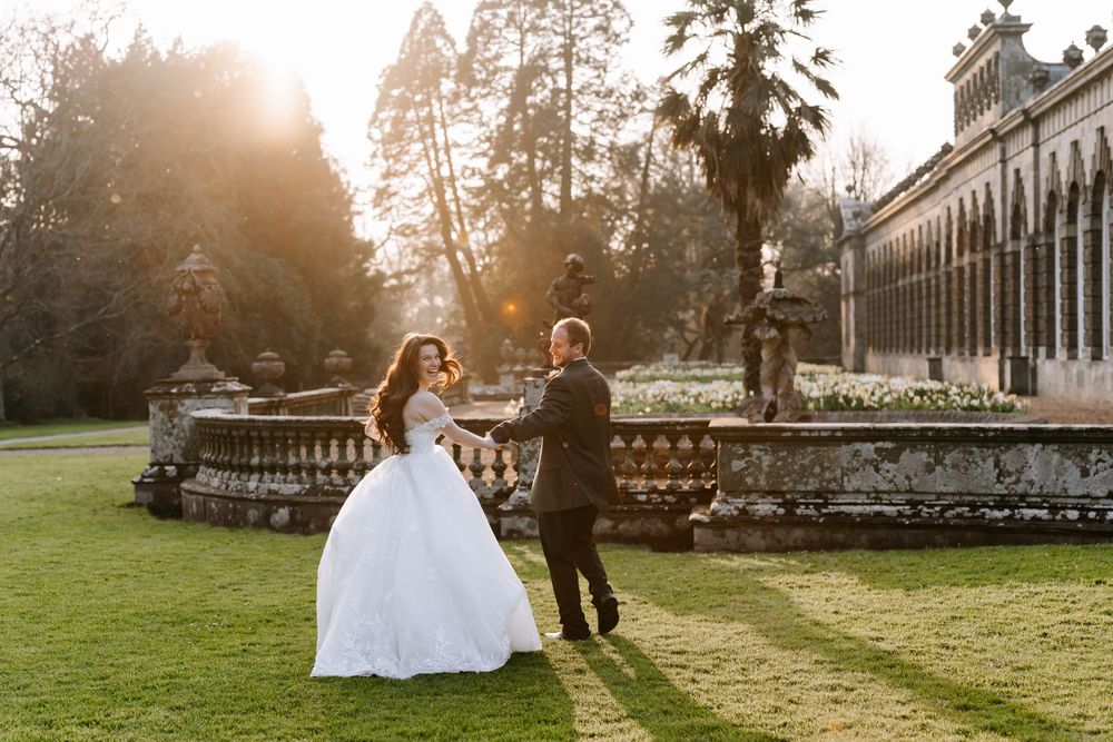 A couple in wedding attire dances on a manicured lawn at sunset near an elegant historic stone building.