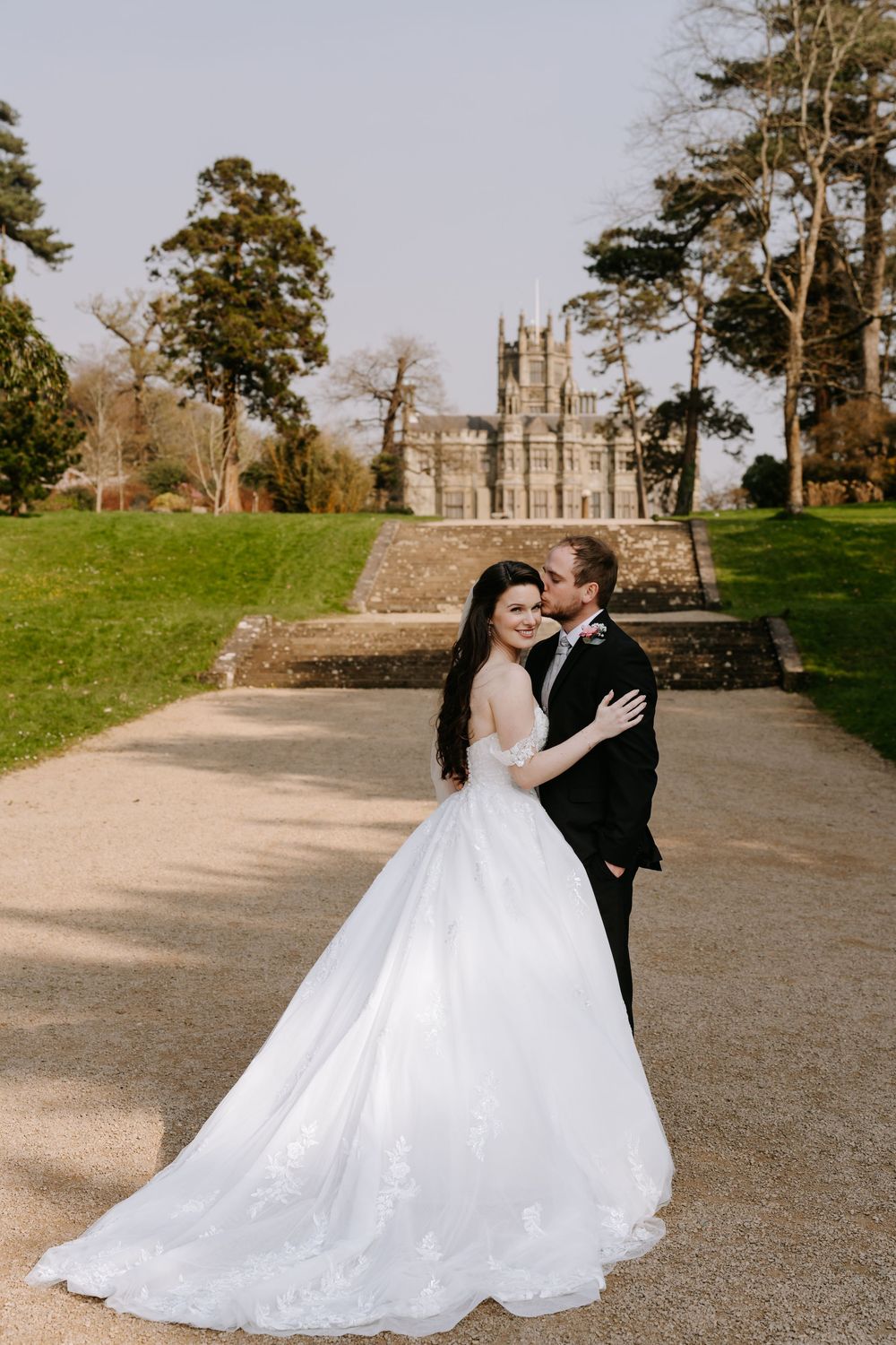 A couple in wedding attire embrace on the grounds of a historic stone building with trees in the background.