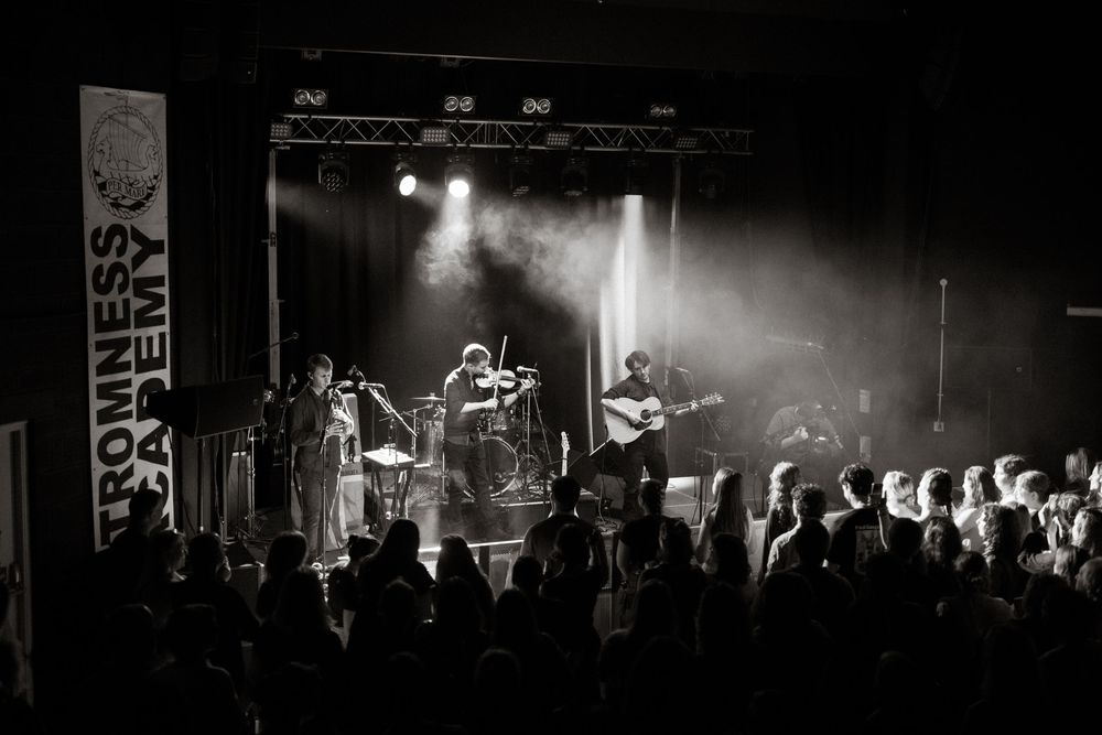 A black and white concert photo showing musicians performing on stage with dramatic lighting and a crowd of silhouettes below.