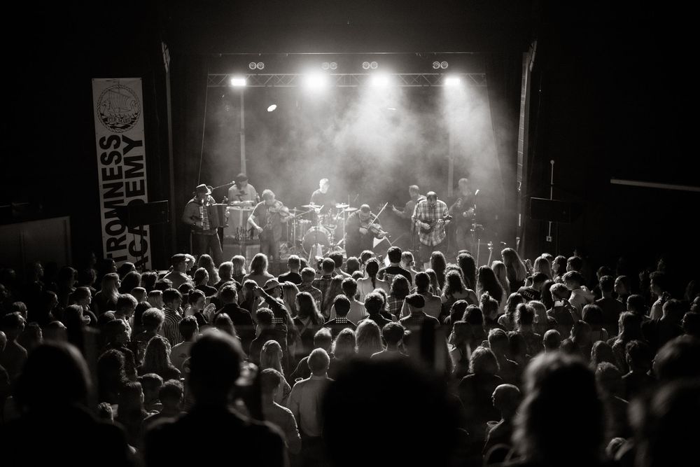 Black and white photo of a crowded concert venue with audience silhouettes and band performing on illuminated stage.