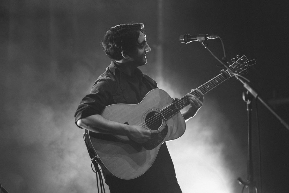 A musician performs on acoustic guitar in black and white silhouette on stage with dramatic back lighting and fog.