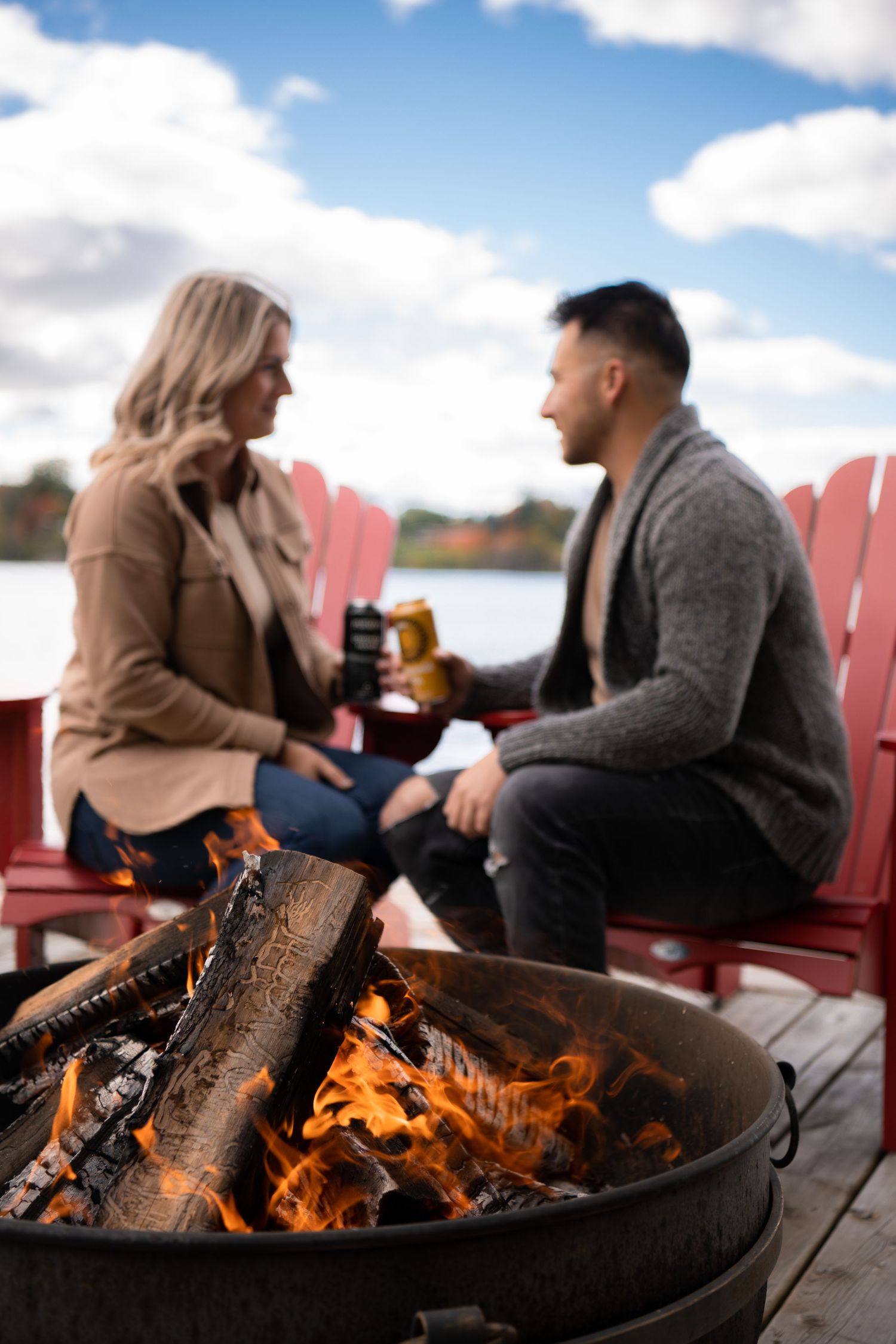 A cozy autumn scene with drinks by a fire pit on red Adirondack chairs near a lake.