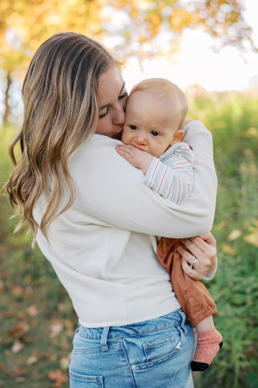 Loving parent in white sweater and jeans embraces baby against autumn backdrop.