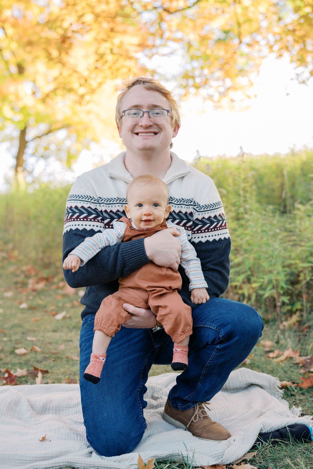 Parent in patterned sweater holds smiling baby during fall outdoor portrait session.