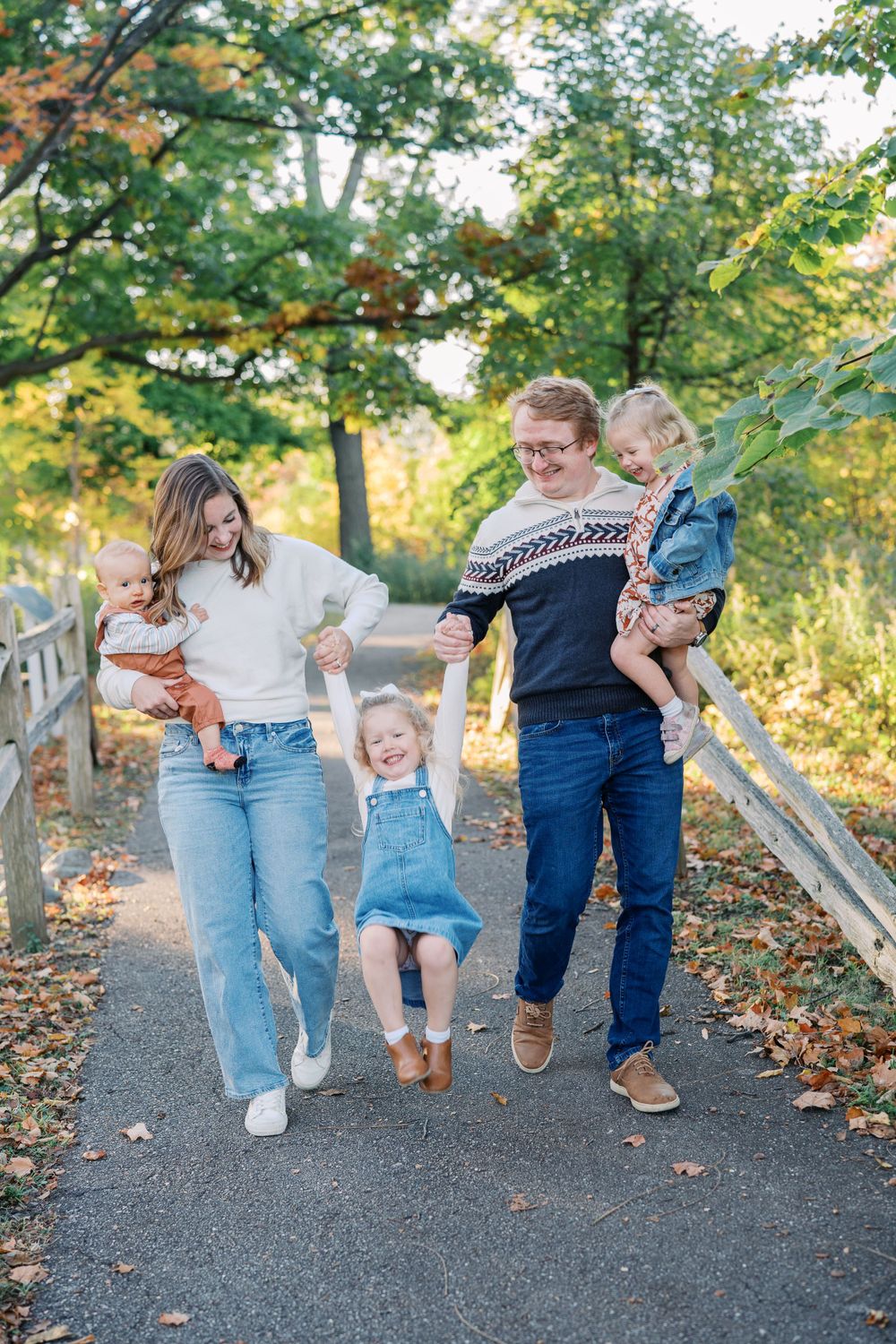Family jumps together holding hands on path surrounded by fall foliage.
