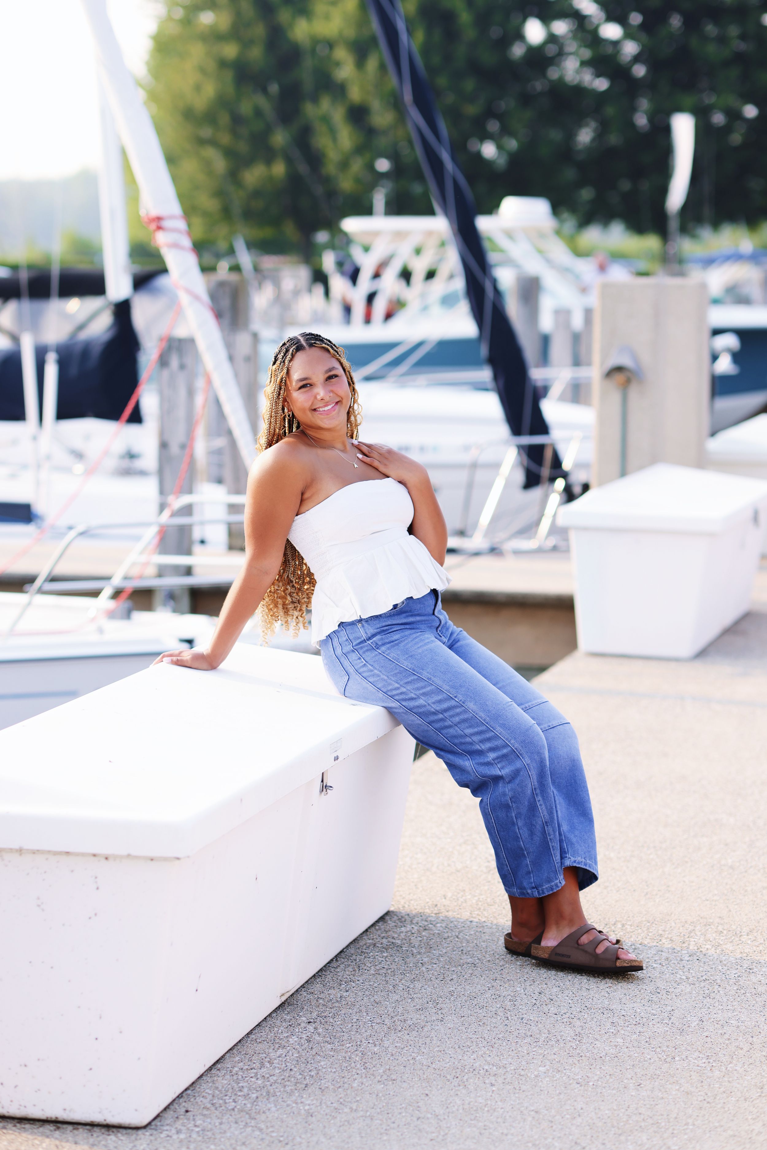 A person in white top and blue jeans poses playfully on white outdoor furniture at a poolside setting.