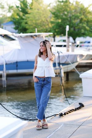 Woman in white top and blue jeans standing on wooden dock near boats at marina on sunny day.