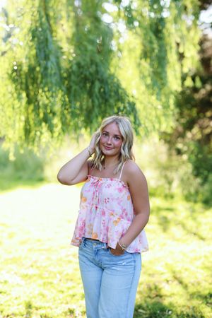 A person in a floral crop top and light jeans poses playfully in front of a weeping willow tree in summer.
