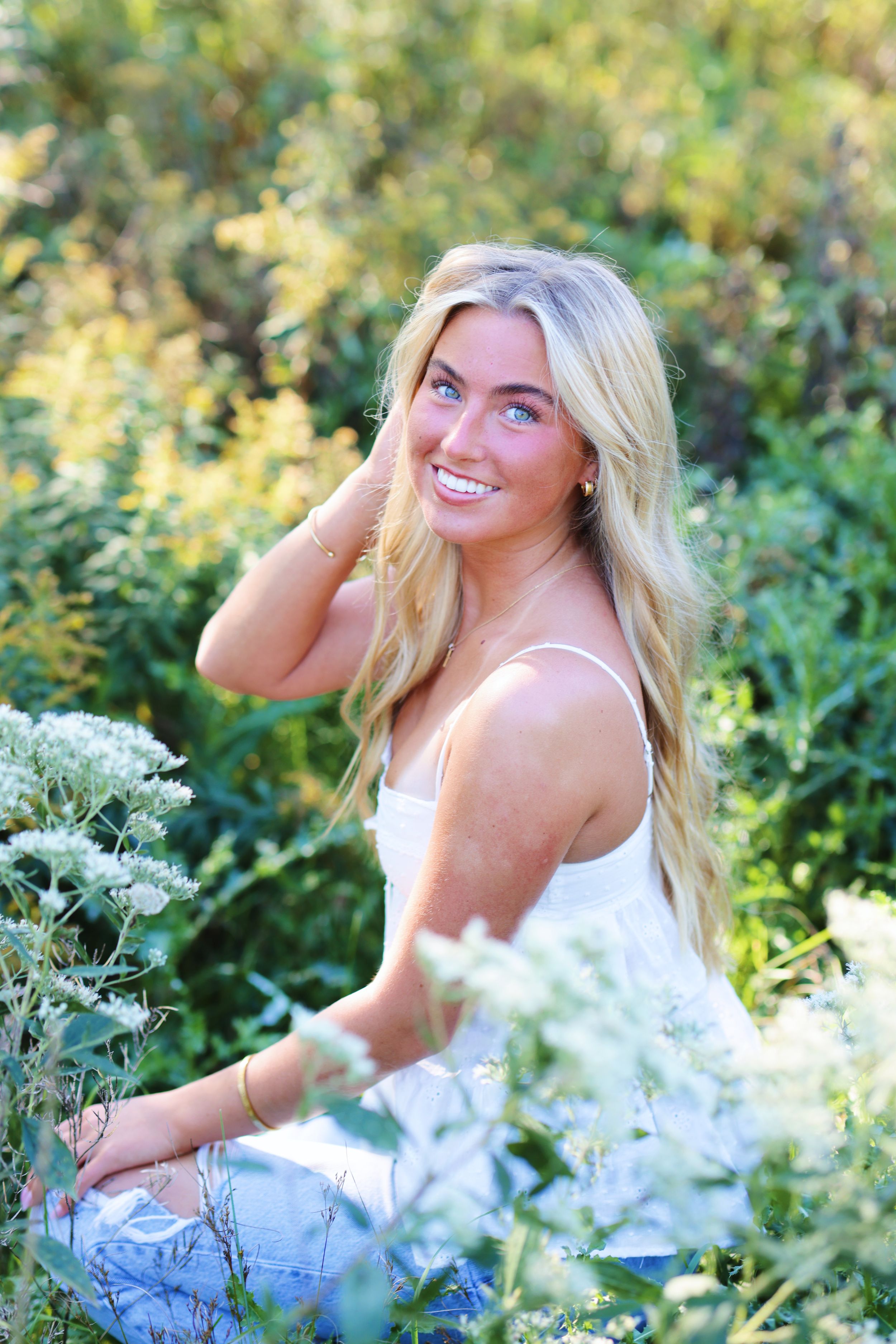 A person in a white dress poses joyfully among wildflowers and greenery in a sunny outdoor setting.
