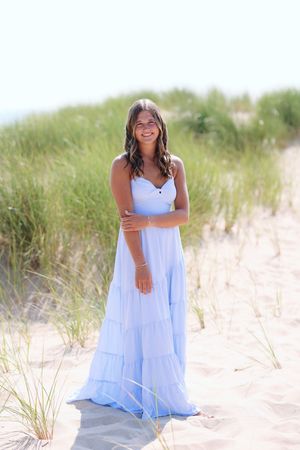 A person in a flowing white dress poses gracefully on sandy beach dunes with coastal grass in the background.
