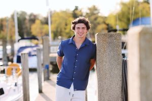 A person stands smiling near a concrete post at a marina on a sunny autumn day.