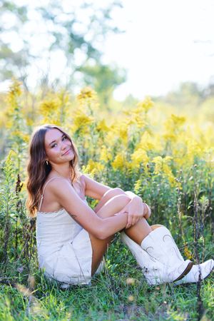 A person in a white dress sits joyfully in a field of blooming yellow wildflowers on a sunny summer day.