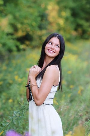 A woman in a white sundress poses joyfully in a lush outdoor setting with green foliage and bokeh lighting effects.
