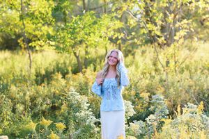 Someone in a light blue top and white skirt stands in a sunlit meadow with wildflowers and trees in the background.