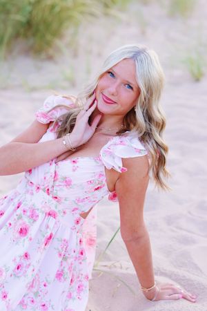 Woman in floral pink and white sundress poses playfully on sandy beach during summer photoshoot.