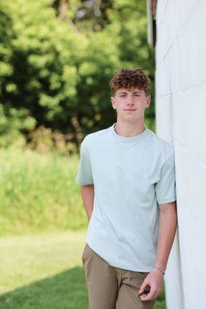 A young person in a light blue t-shirt and khaki pants stands casually against a white wall outdoors on a sunny day.