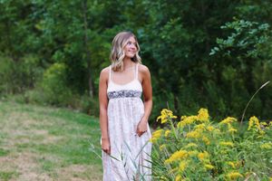 A woman in a white summer dress stands in a field next to blooming yellow wildflowers on a sunny day.