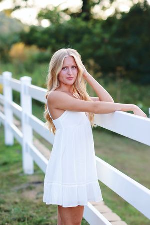 A person in a white summer dress poses playfully along a white wooden fence in a sunny outdoor setting.