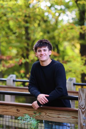 A person in a black sweater leans on a wooden railing outdoors with green foliage in the background.