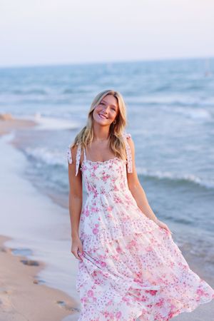 A person in a flowing floral summer dress walks along a peaceful beach at sunset with waves in the background.
