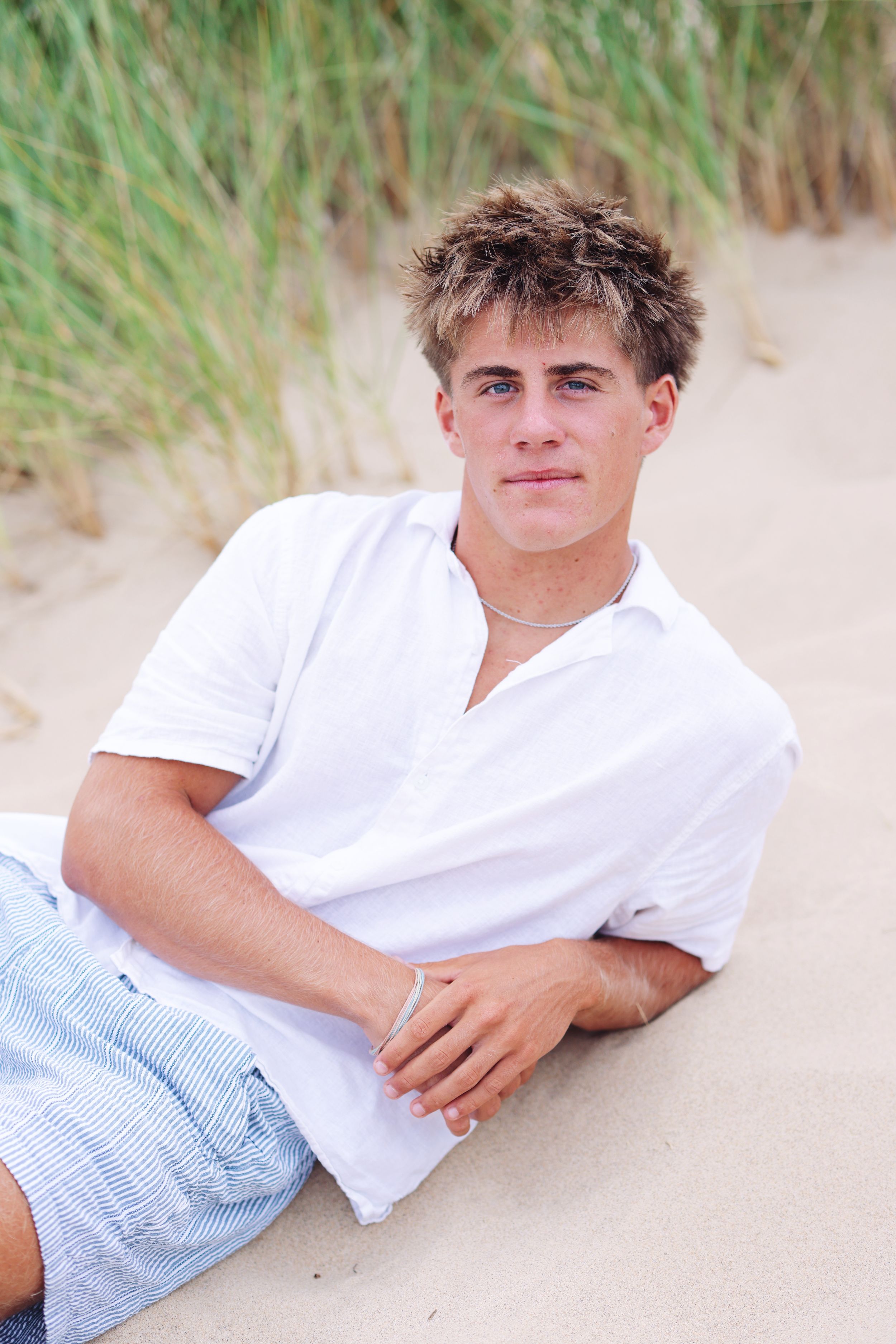 Young person in white shirt and blue shorts relaxing on sandy beach with grass in background.