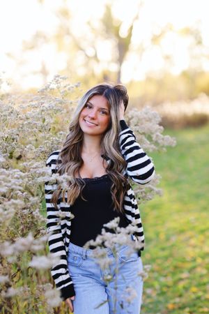 A person in a striped cardigan and jeans poses outdoors among delicate wildflowers on a sunny autumn day.