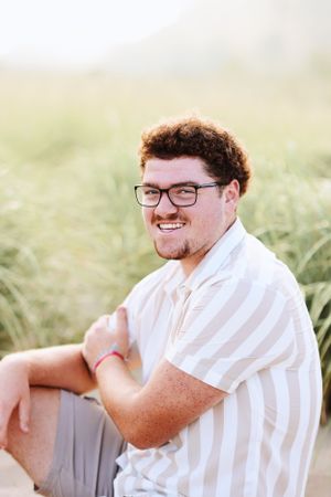 A cheerful person wearing glasses and a white striped button-down shirt sits outdoors in a grassy field during golden hour.