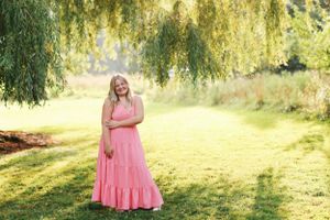 A figure in a flowing pink maxi dress stands on a sunlit lawn beneath drooping willow tree branches.