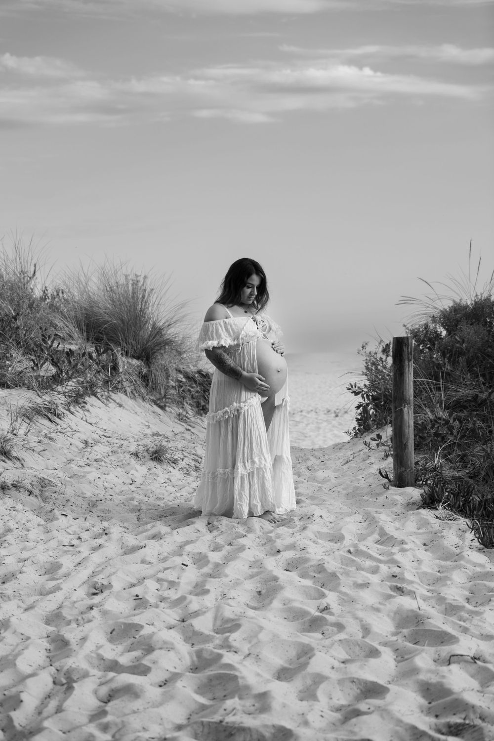 Expectant mother in white dress standing on sandy dunes in black and white photography.