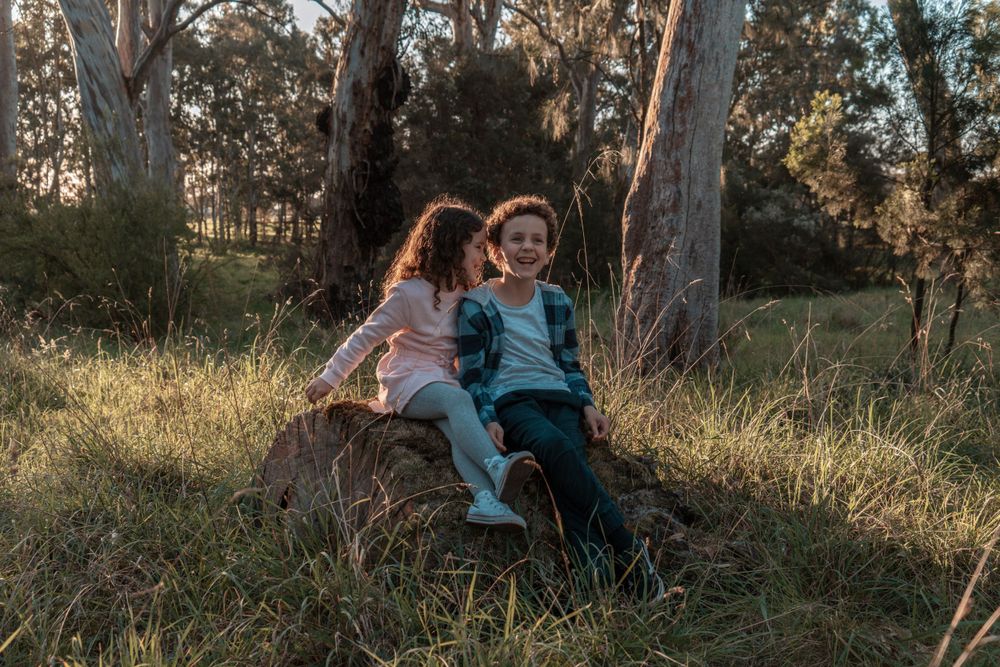 Two children sitting together and laughing on a log at sunset in a forest setting.