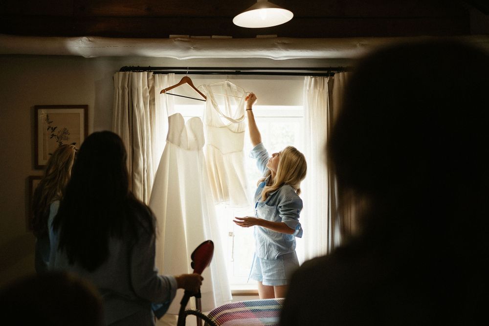 A white wedding dress hangs illuminated in a window as silhouettes and figures are visible in the dimly lit room.