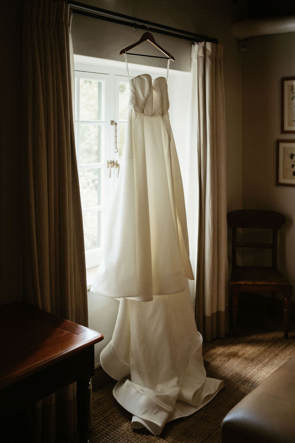 A white wedding dress hangs elegantly by a window in a rustic room with natural lighting and wooden floors.