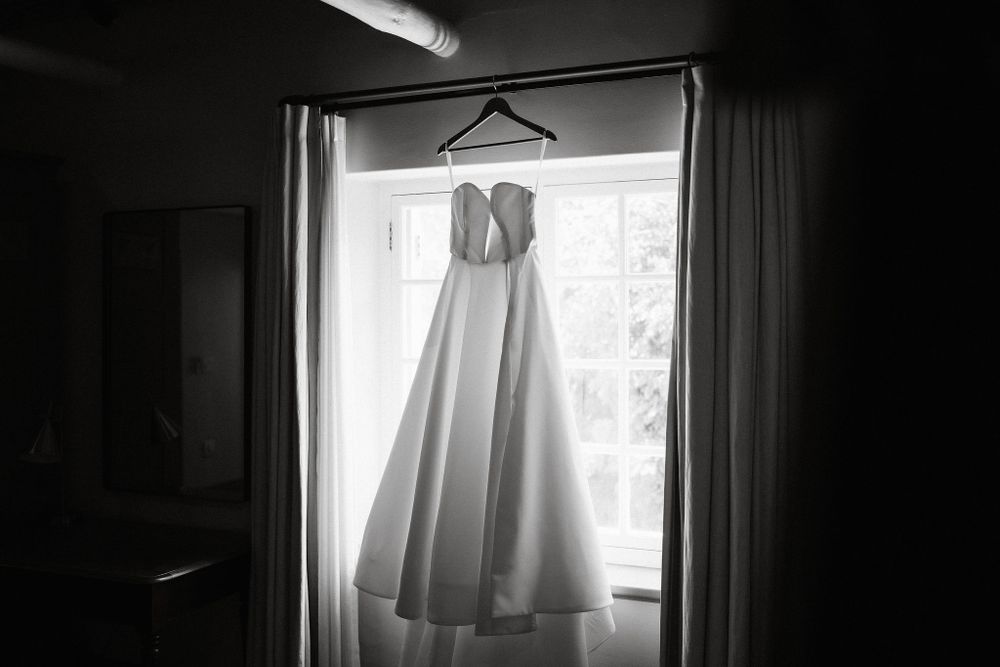 A white wedding dress hangs silhouetted against a bright window in a dark room creating a dramatic contrast.