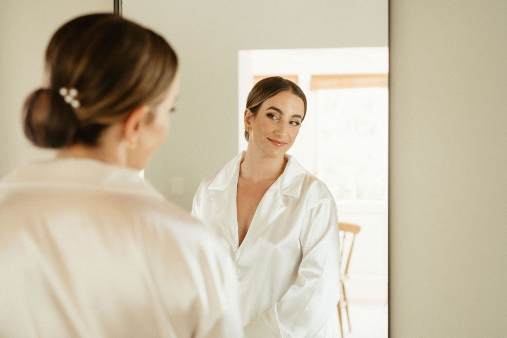 Woman in white robe looking at her reflection in mirror during quiet morning moment.