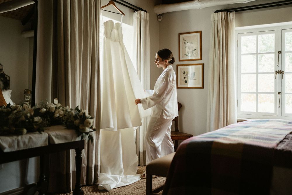 Someone in white preparing for a wedding stands next to an elegant white gown hanging by a bright window in a bedroom.