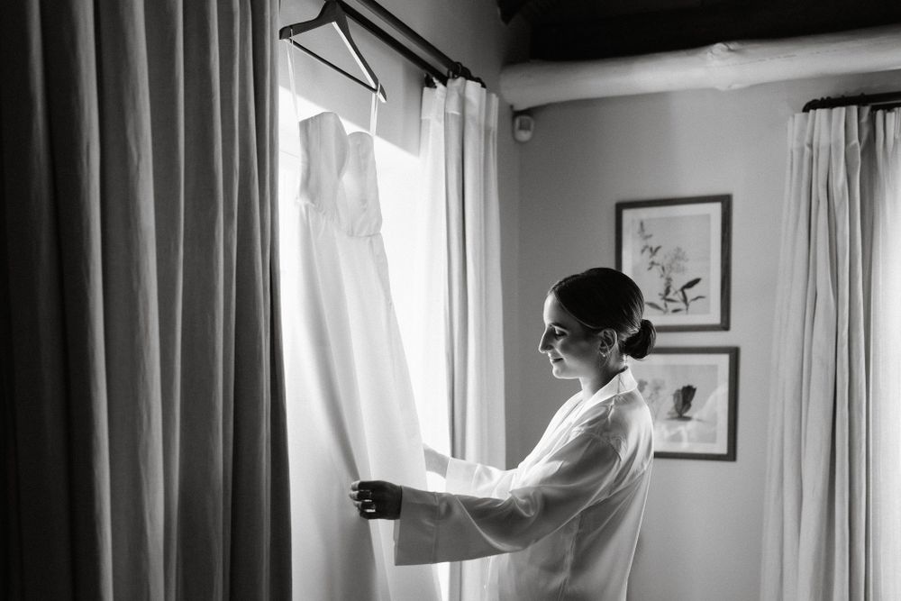 A white wedding dress hangs by a window as someone in a robe gazes at it during morning preparations.