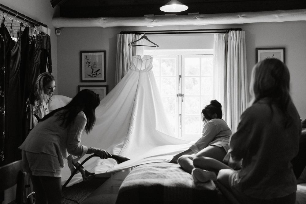 Black and white photo showing people steaming and preparing a wedding dress hanging in front of a window.