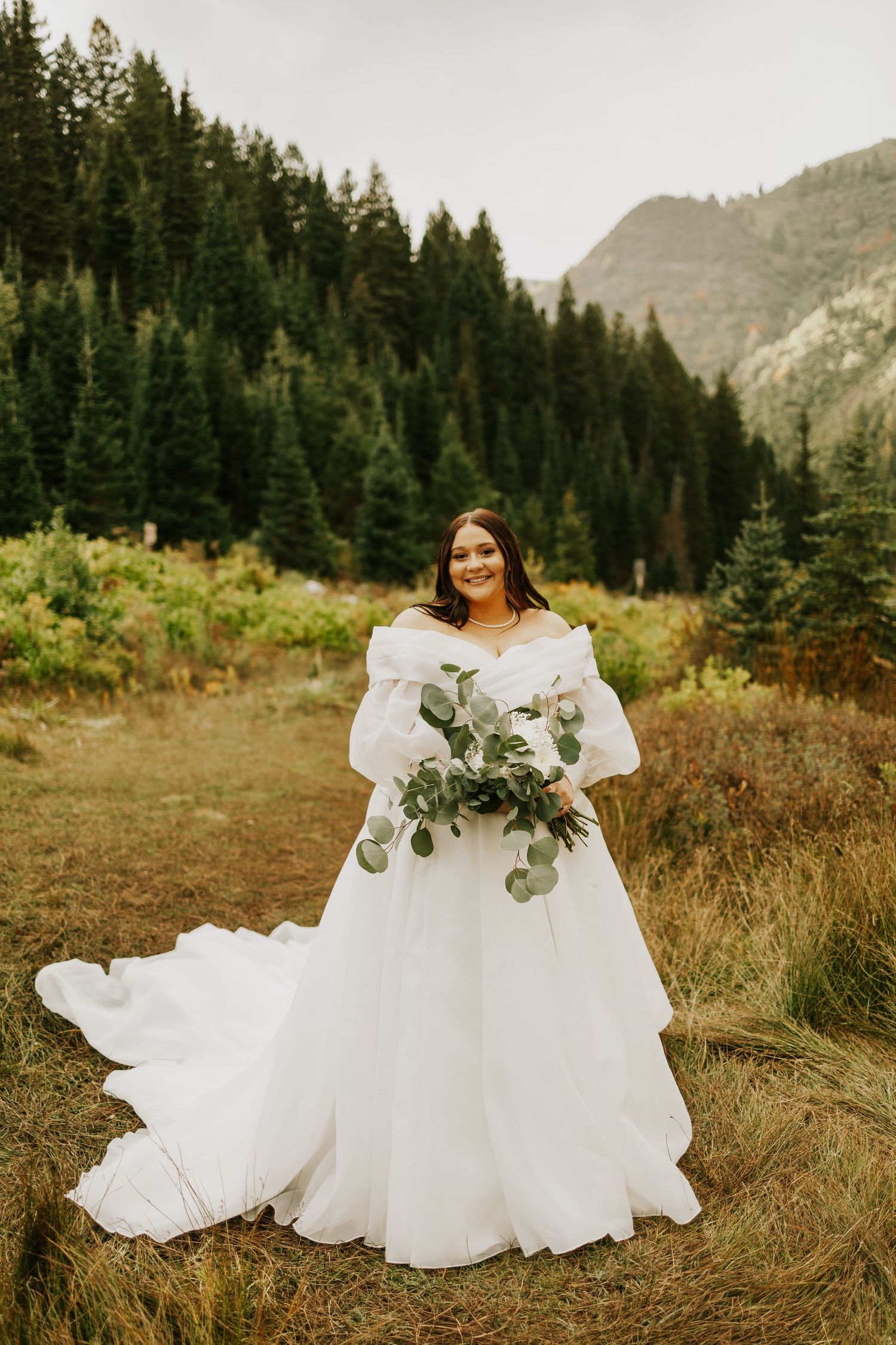 Bride in off-shoulder white ball gown holding eucalyptus bouquet in mountain forest setting.