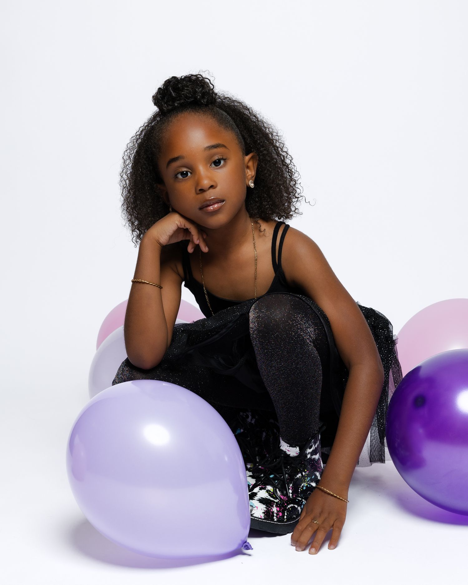 A child in a black outfit poses with lavender and purple balloons against a white background for a birthday portrait session.