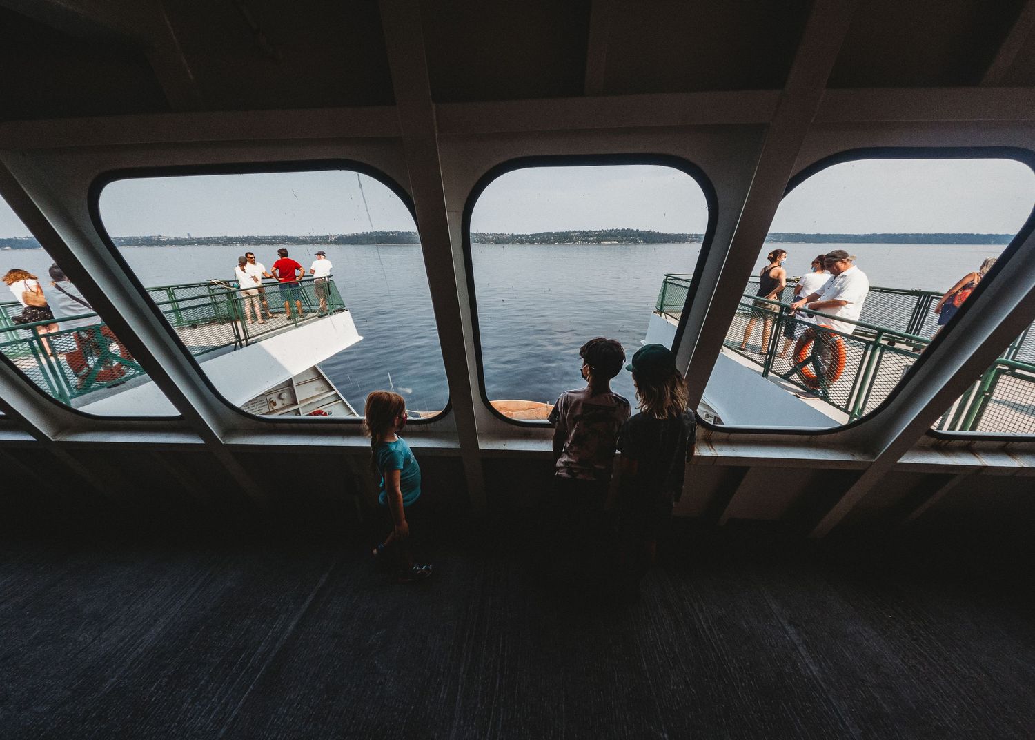 Passengers look out through large ferry windows at the ocean view during a boat journey.