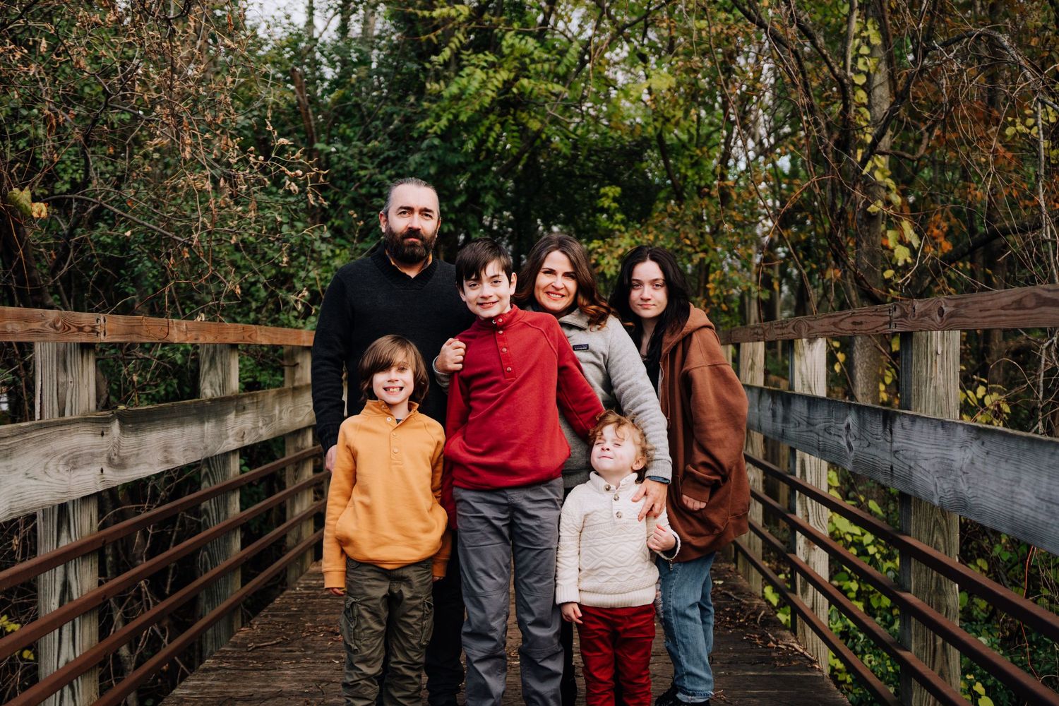 Family of five standing together on wooden bridge surrounded by trees.