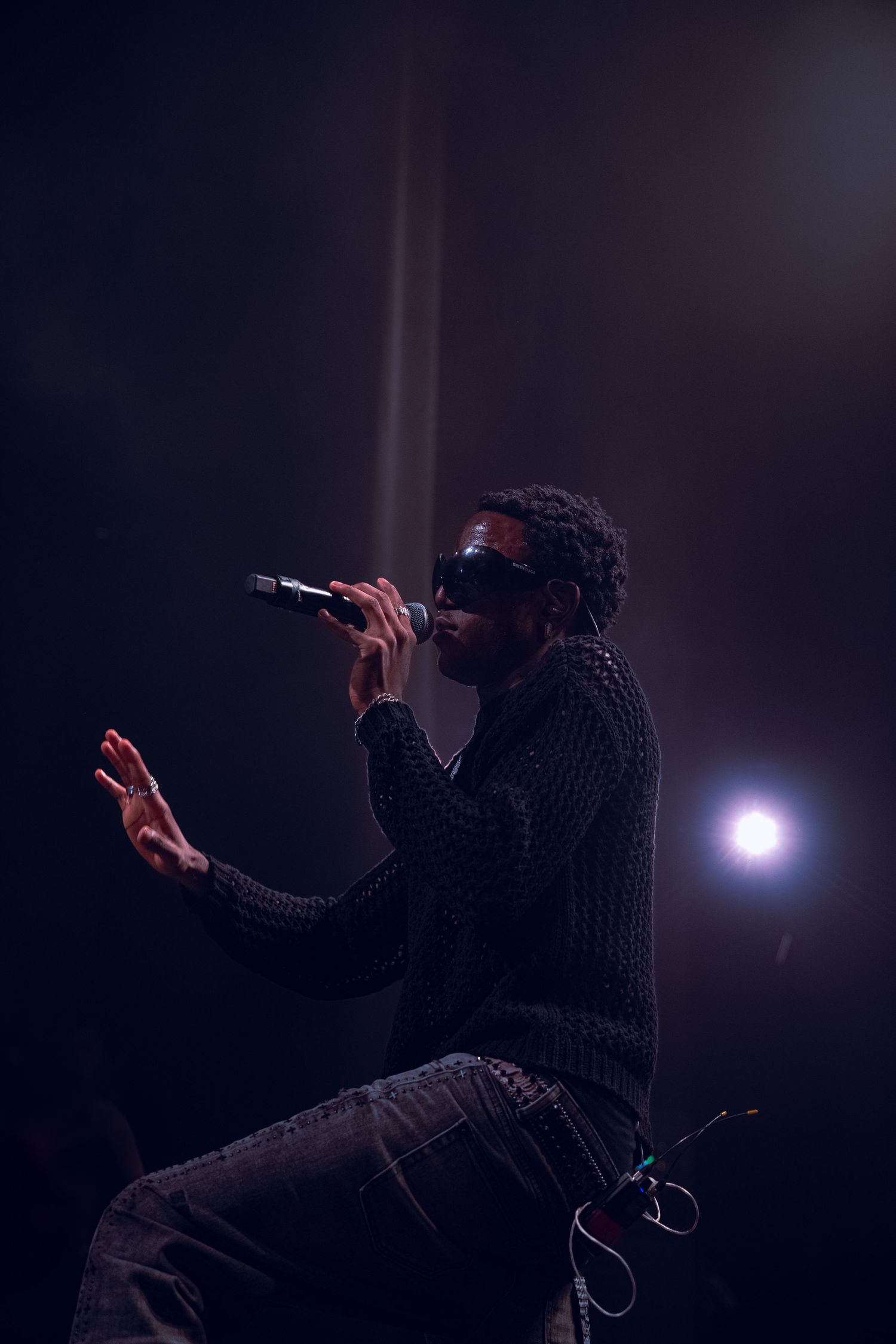 Silhouetted performer in sunglasses sings passionately on dark stage with dramatic lighting and beam effect.