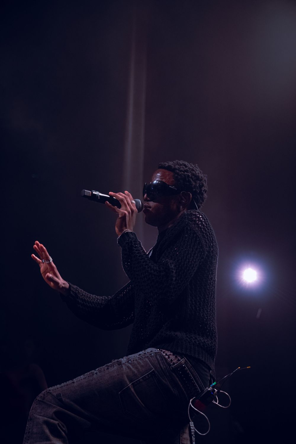 Silhouetted performer in sunglasses sings passionately on dark stage with dramatic lighting and beam effect.