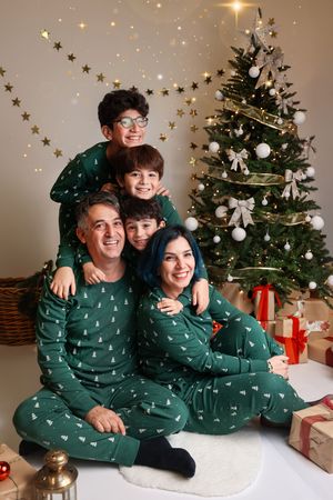 A family wearing matching green holiday pajamas poses together by a decorated Christmas tree with wrapped presents.