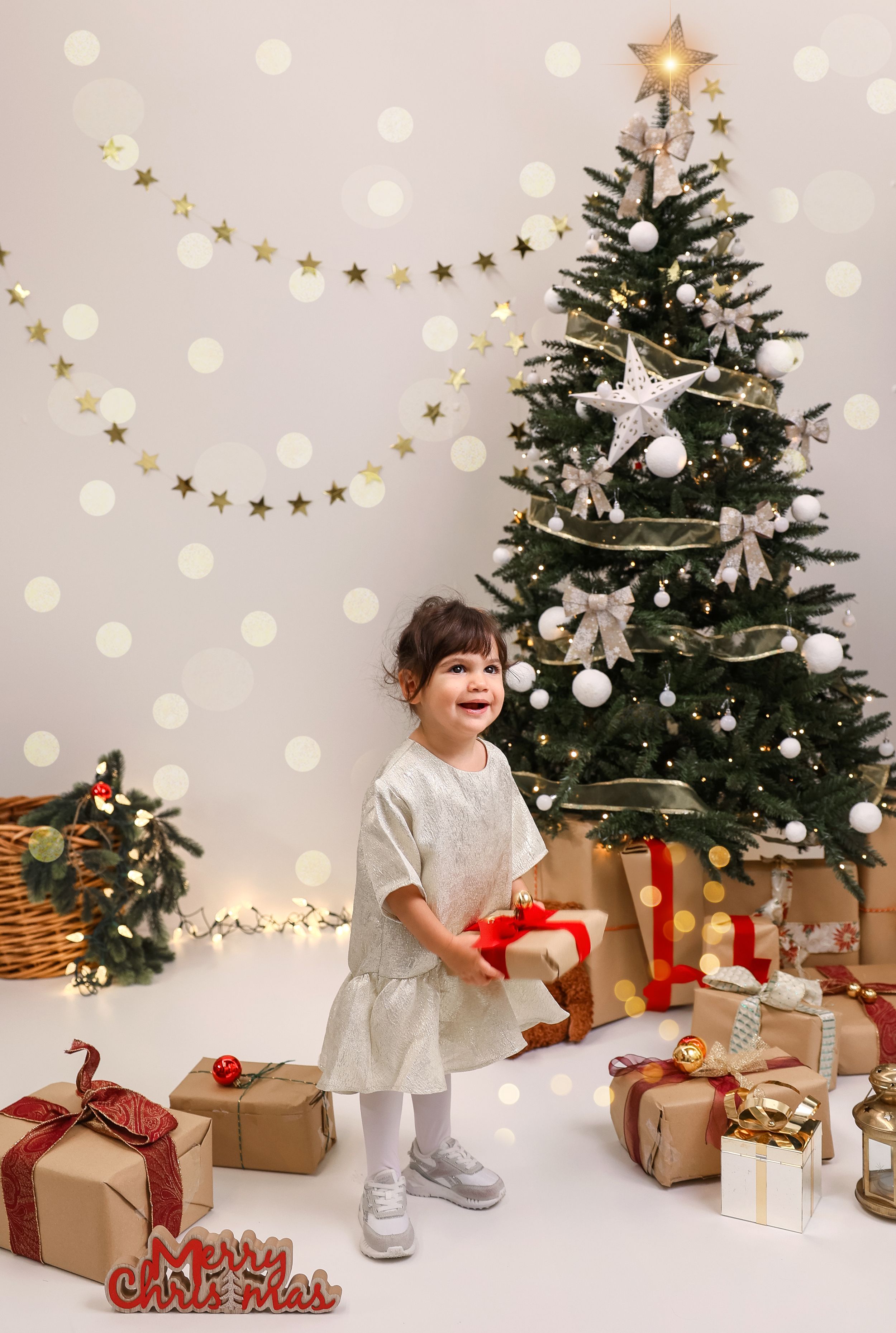 Decorated Christmas tree with white and gold ornaments stands beside wrapped presents and star garland on polka dot background.