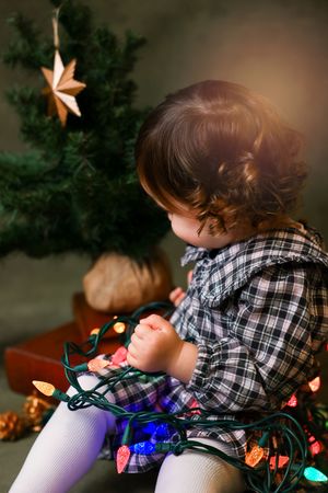 A child in a plaid shirt lies on their side while tangled in colorful Christmas lights.