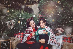 Two people in festive Christmas sweaters share hot cocoa surrounded by holiday decorations and twinkling lights.