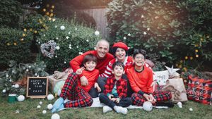 Holiday family portrait with matching red and plaid outfits sitting on white blanket with festive decorations.